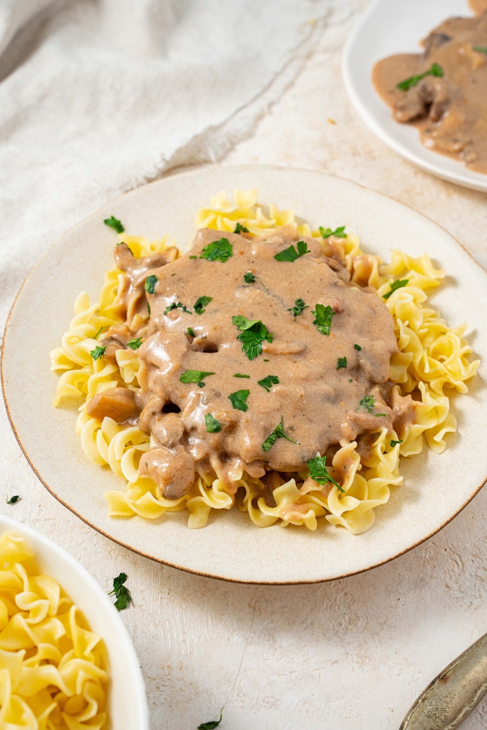 A plate filled with crock pot cubed steak and gravy on a bed of egg noodles with fresh parsley on top. There is an additional plate of egg noodles and an additional plate of cube steak in gravy on either side.