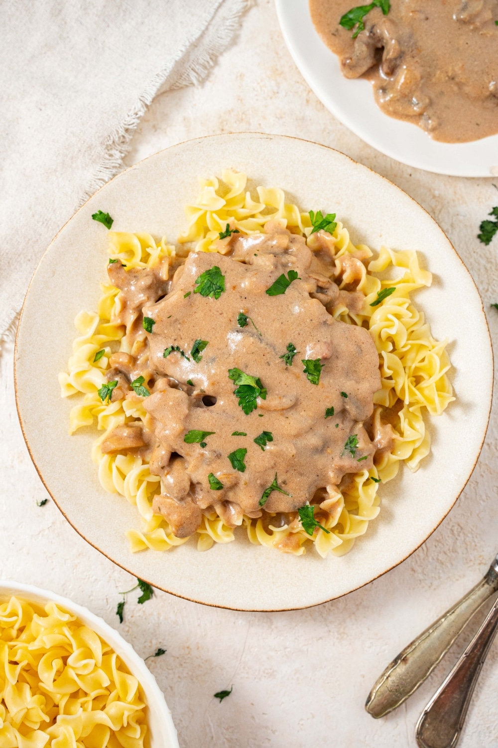 An overhead shot of a plate filled with crock pot cubed steak and gravy on a bed of egg noodles with fresh parsley on top. There is an additional plate of egg noodles and an additional plate of cube steak in gravy on either side.