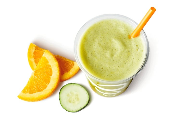 A cup of a cucumber smoothie with two orange slices and cucumber slices next to it.