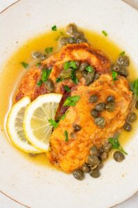 A plate of two fried calamari steaks with a lemon caper sauce. The plate is garnished with sliced lemon and fresh parsley. There is a bowl of capers next to the plate.