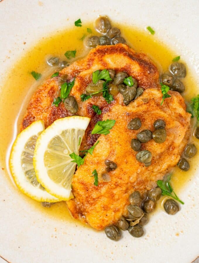 A plate of two fried calamari steaks with a lemon caper sauce. The plate is garnished with sliced lemon and fresh parsley. There is a bowl of capers next to the plate.