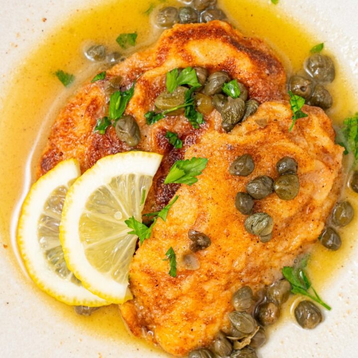 A plate of two fried calamari steaks with a lemon caper sauce. The plate is garnished with sliced lemon and fresh parsley. There is a bowl of capers next to the plate.