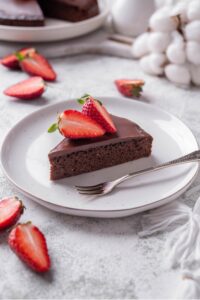 A slice of chocolate protein cake topped with sliced strawberries on a white ceramic plate. There is a fork next to the slice.