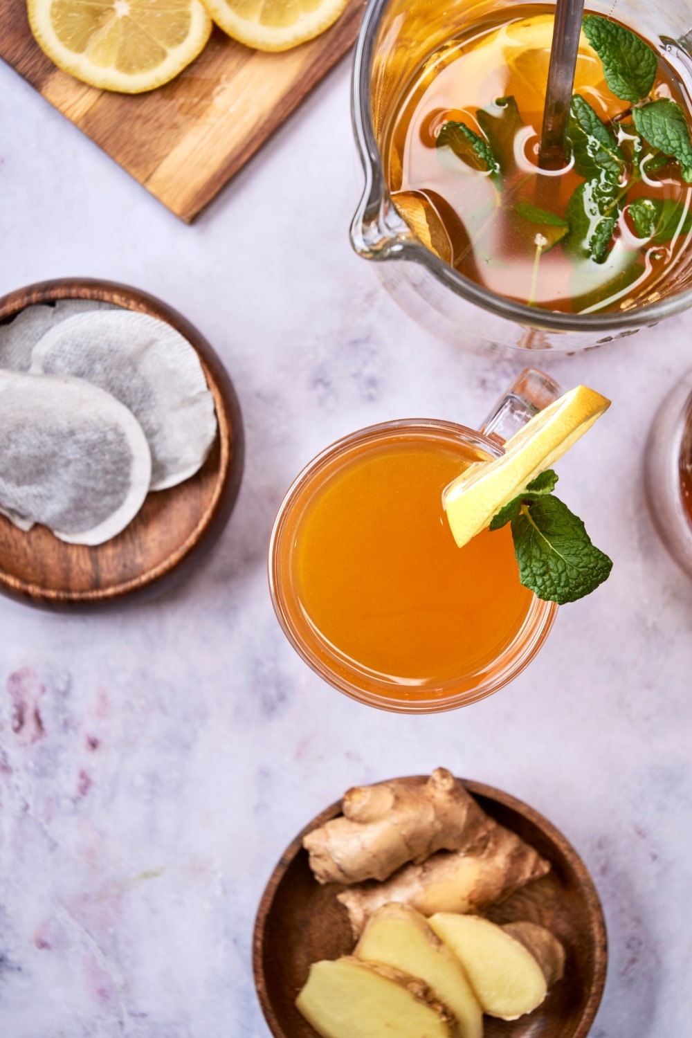 An overhead shot of flu bomb tea in a glass mug garnished with fresh mint and a slice of lemon. There is a pitcher of flu bomb tea next to it, along with two wooden bowls containing fresh ginger and green tea bags.