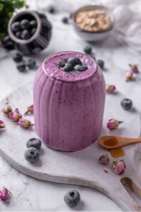 A ribbed glass filled with a high fiber smoothie served on a wooden board. The smoothie is topped with blueberries and chia seeds and there are blueberries and dried flower buds scattered around the board.
