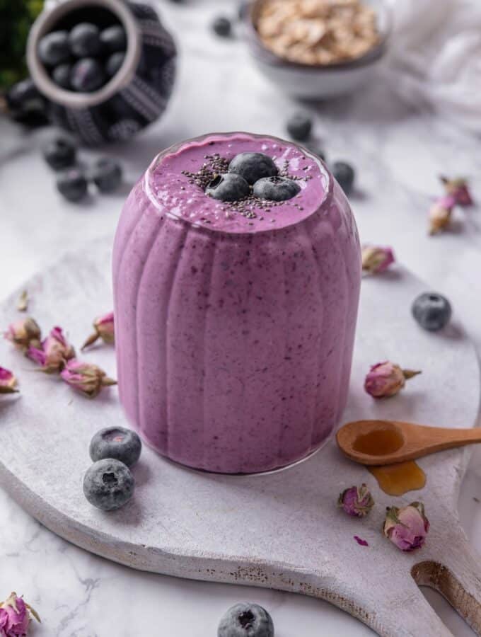 A ribbed glass filled with a high fiber smoothie served on a wooden board. The smoothie is topped with blueberries and chia seeds and there are blueberries and dried flower buds scattered around the board.