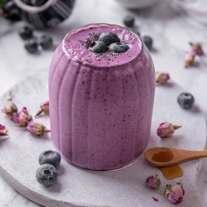 A ribbed glass filled with a high fiber smoothie served on a wooden board. The smoothie is topped with blueberries and chia seeds and there are blueberries and dried flower buds scattered around the board.