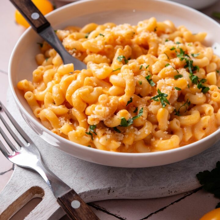 A bowl of protein mac and cheese on a white wooden server. There is a fork inserted into the macaroni and another fork next to the bowl. There is a second bowl of mac and cheese in the background.