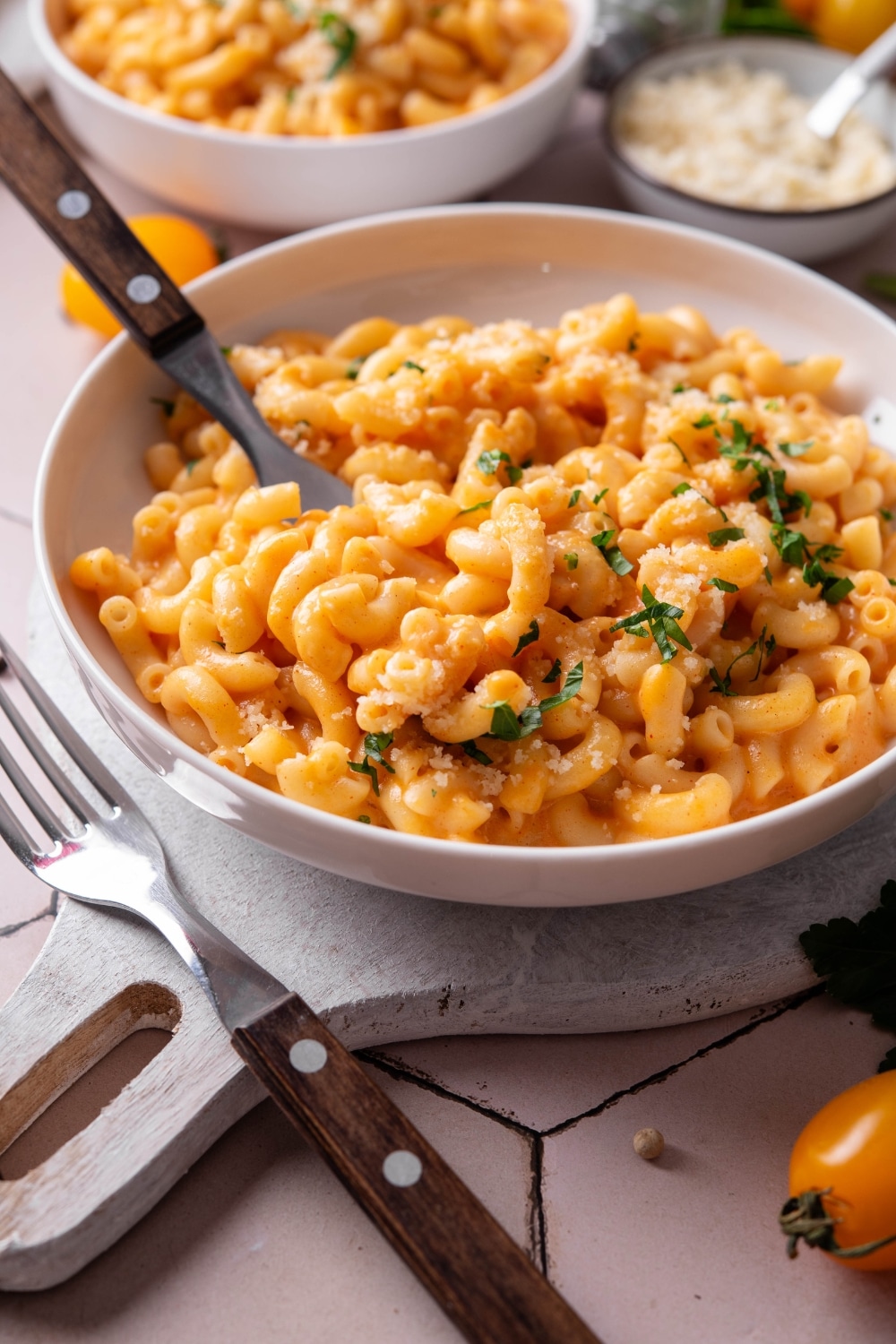 A bowl of protein mac and cheese on a white wooden server. There is a fork inserted into the macaroni and another fork next to the bowl. There is a second bowl of mac and cheese in the background.