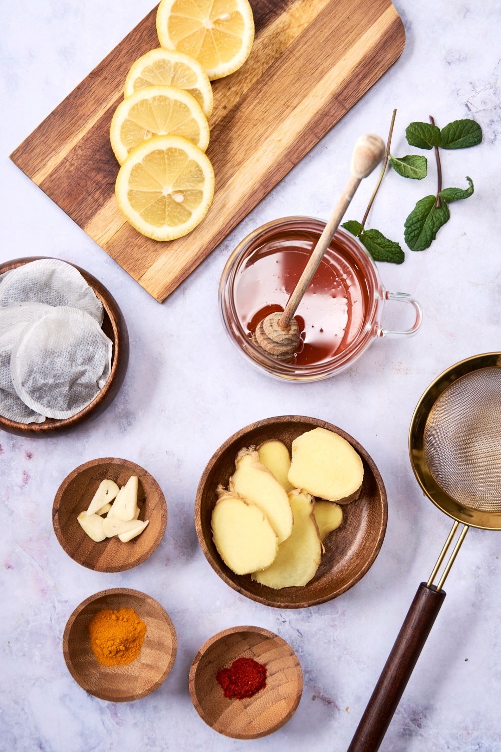 An overhead shot of several bowls containing various ingredients for flu bomb tea, including freh ginger, honey, green tea bags, garlic, turmeric, and ground chili peppers. There is also a small strainer and a wooden board with sliced lemons next to the bowls.