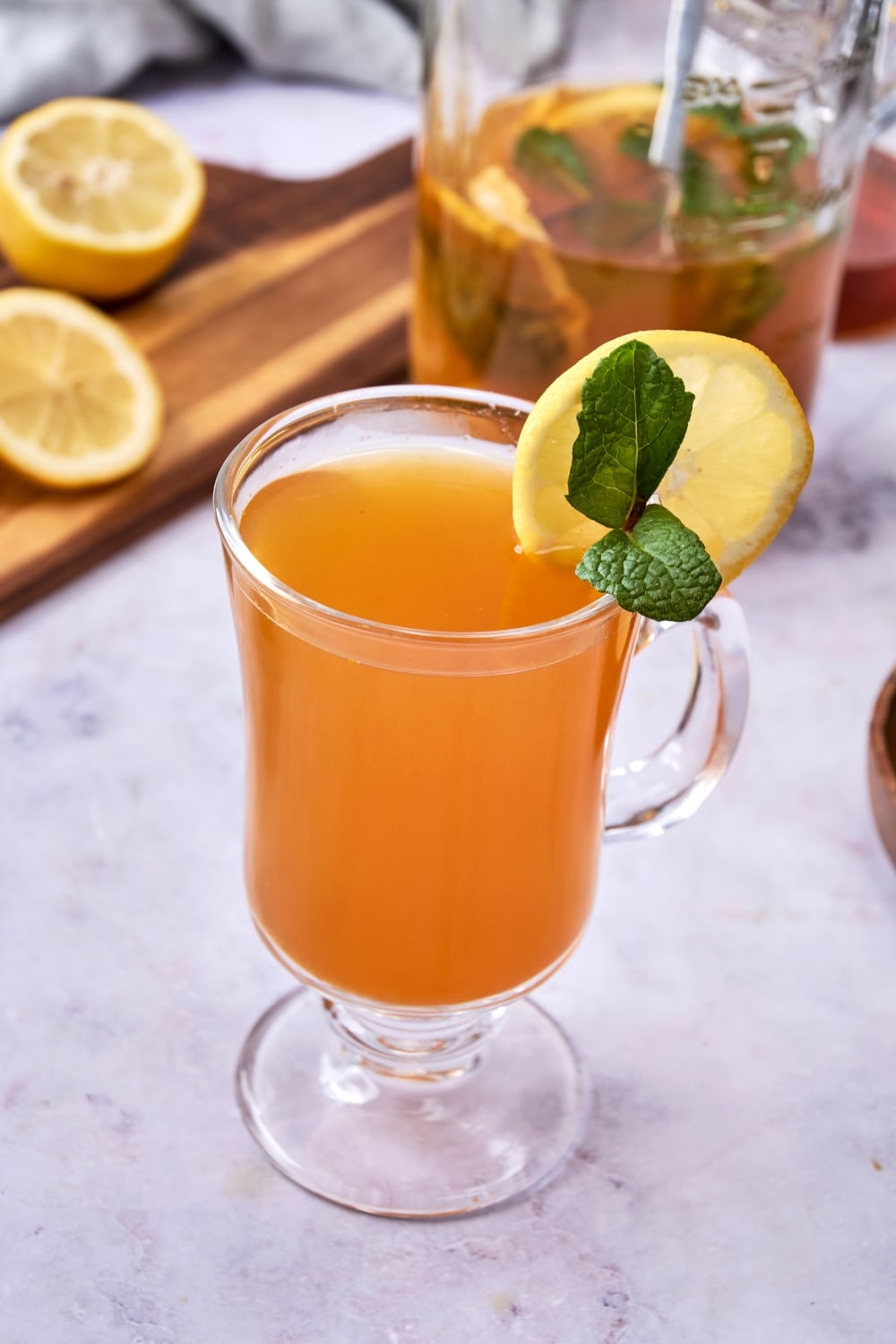 A glass tea mug filled with flu bomb tea garnished with fresh mint and a slice of lemon. There is a glass pitcher of flu bomb tea and a wooden board with sliced lemon in the background.