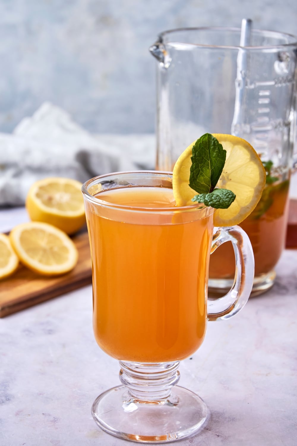 A glass tea mug filled with flu bomb tea garnished with fresh mint and a slice of lemon. There is a glass pitcher of flu bomb tea and a wooden board with sliced lemon in the background.