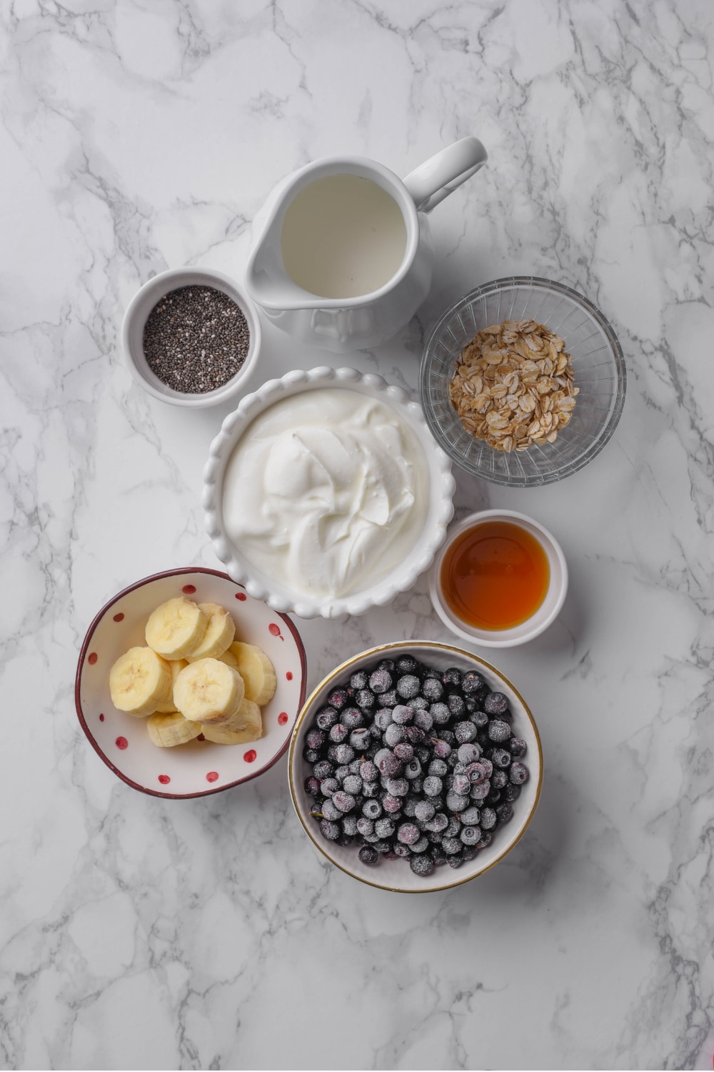 An overhead shot of several bowls of various shapes and sizes containing the ingredients to make a high fiber smoothie including frozen blueberries, oats, sliced banana, chia seeds, greek yogurt, and milk.