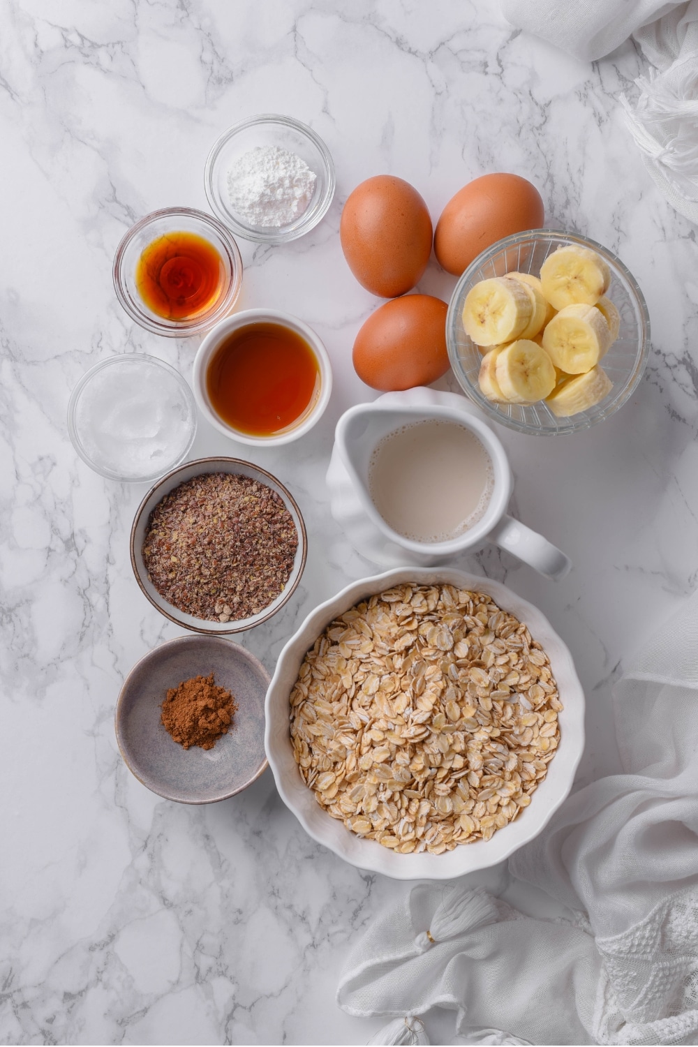 An overhead shot of several bowls of various sizes containing ingredients for high fiber pancakes such as oats, sliced banana, flax seeds, cinnamon, almond milk, vanilla, syrup, and eggs.