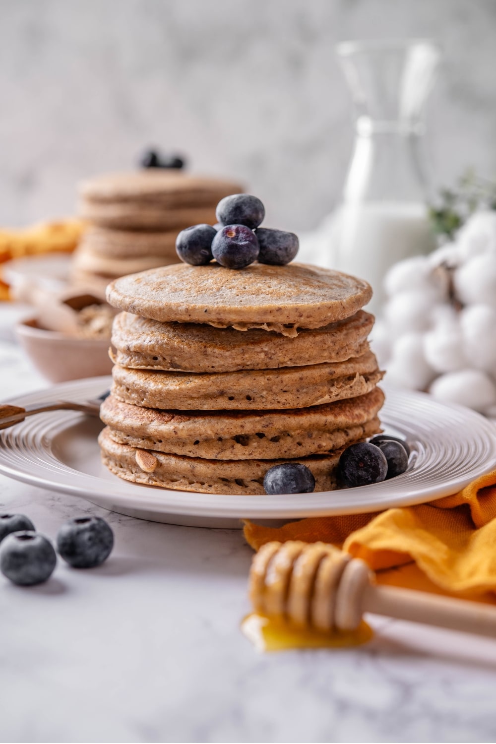 A stack of five high fiber pancakes on a white ceramic plate. There is a fork to the side of the stack and the pancakes are topped with blueberries with extra blueberries scattered on the plate.