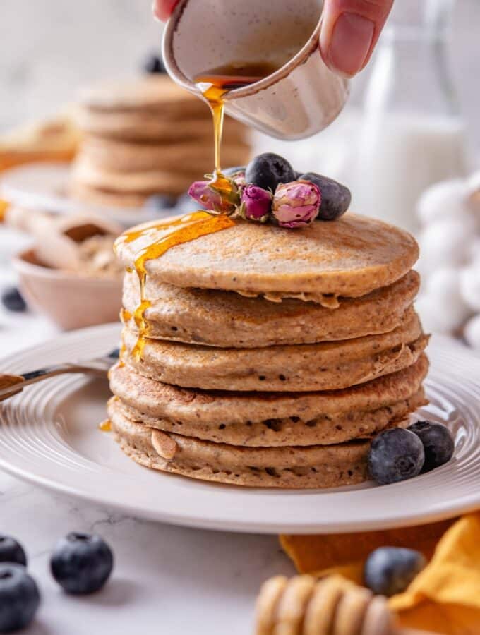 A stack of five high fiber pancakes on a white ceramic plate. The pancakes are topped with blueberries and dried flower buds and there is a hand pouring maple syrup from a ceramic ramekin over the pancakes.