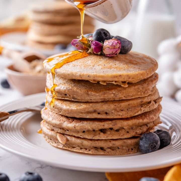 A stack of five high fiber pancakes on a white ceramic plate. The pancakes are topped with blueberries and dried flower buds and there is a hand pouring maple syrup from a ceramic ramekin over the pancakes.