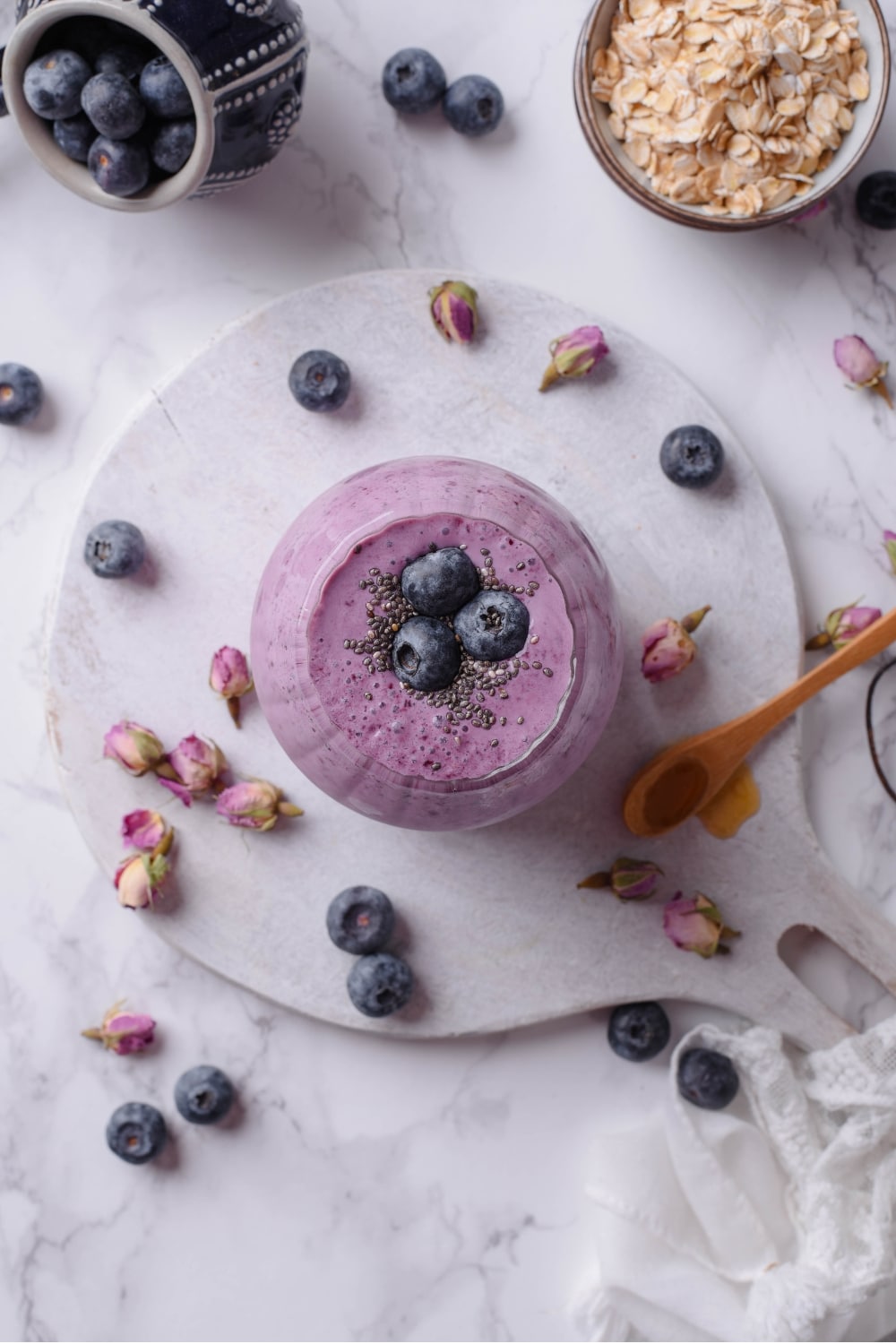 An overhead shot of a glass filled with a high fiber smoothie served on a wooden board. The smoothie is topped with blueberries and chia seeds and there are blueberries and dried flower buds scattered around the board. There is a small bowl of oats and a small bowl of blueberries off to the side.