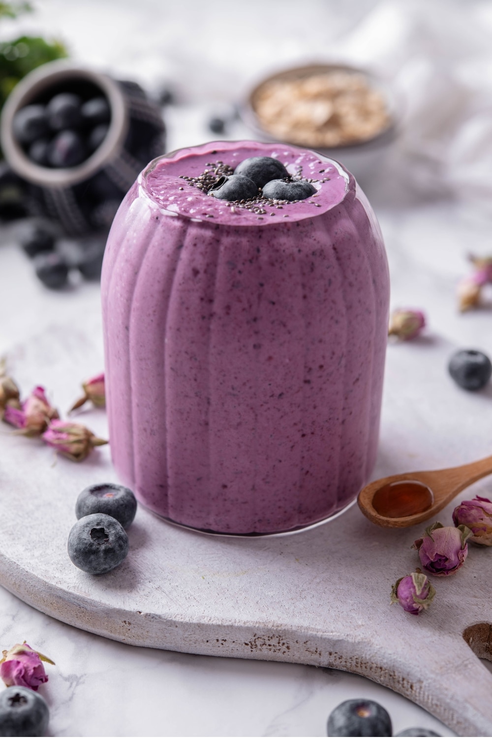 A ribbed glass filled with a high fiber smoothie served on a wooden board. The smoothie is topped with blueberries and chia seeds and there are blueberries and dried flower buds scattered around the board.