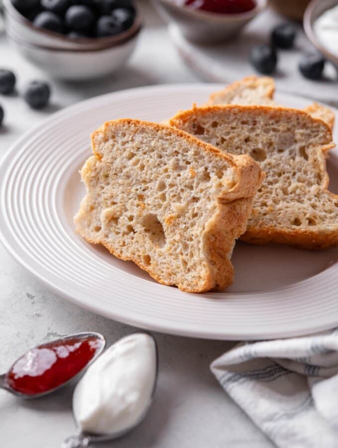 Two slices of protein bread stacked upright on a white plate. There is a napkin next to the plate along with two spoons filled with jam and yogurt.