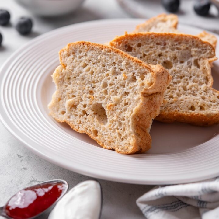 Two slices of protein bread stacked upright on a white plate. There is a napkin next to the plate along with two spoons filled with jam and yogurt.