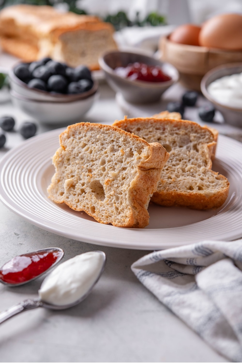 Two slices of protein bread stacked upright on a white plate. There is a napkin next to the plate and an additional loaf in the background.