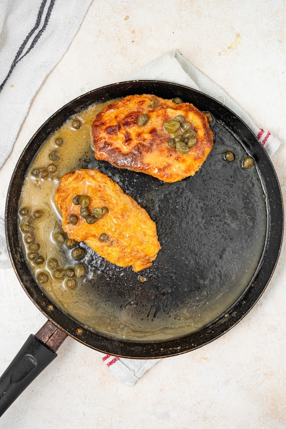 An overhead shot of a large skillet with two fried calamari steaks basting in a lemon caper sauce.