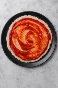 An overhead shot of an uncooked pizza dough on a baking pizza pan. The dough is topped with pizza sauce.