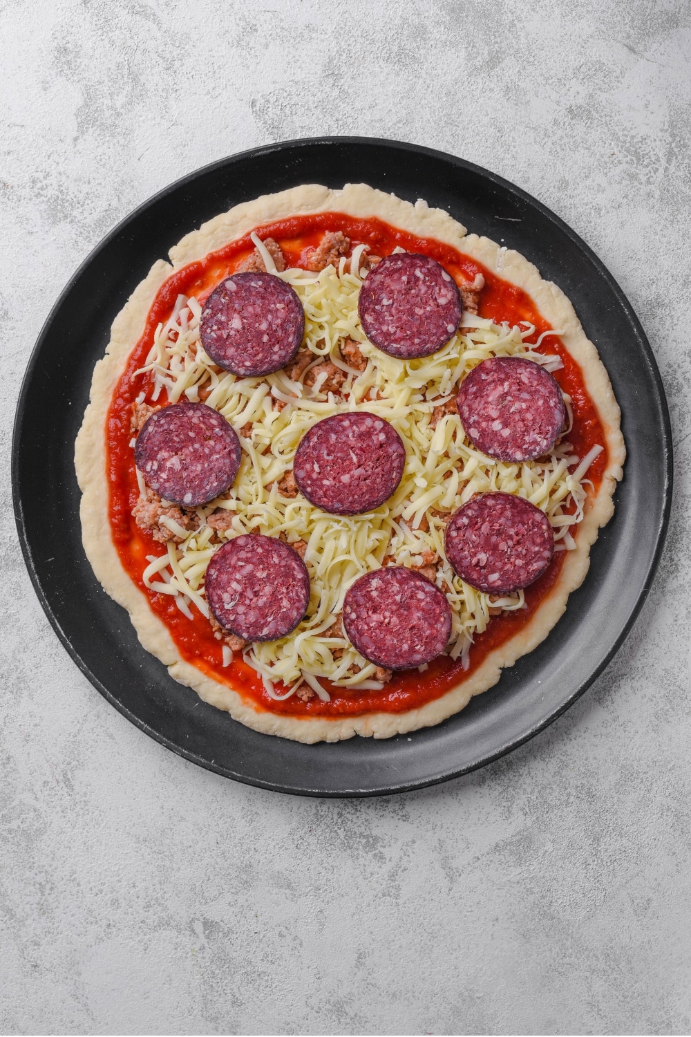 An overhead shot of an uncooked protein pizza on a baking pizza pan. The pizza is topped with sauce, shredded mozzarella cheese, pepperoni, and sausage.