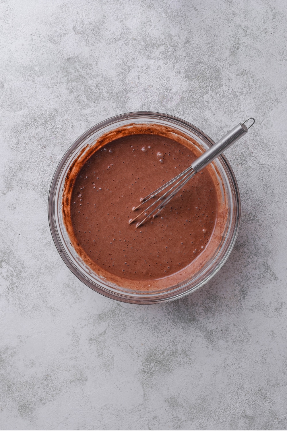 An overhead shot of a glass bowl filled with chocolate protein cake batter. There is a whisk in the bowl.