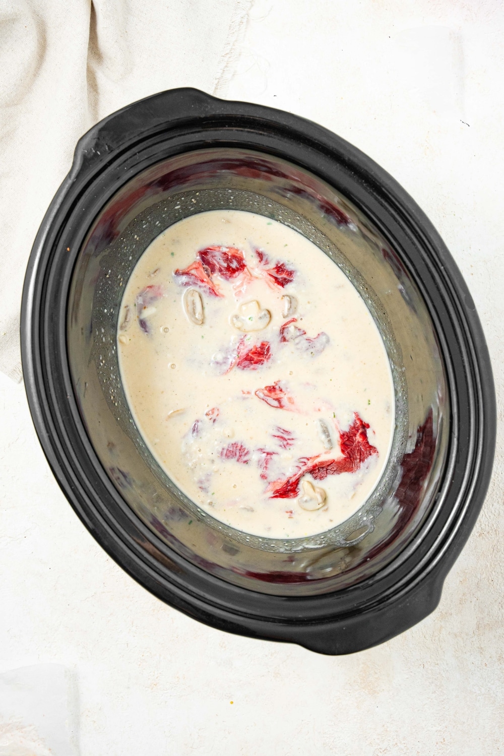 An overhead shot of a slow cooker filled with a beef broth and cream of mushroom soup mixture, with cube steaks submerged in the sauce.