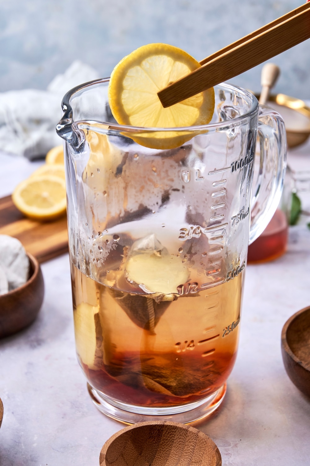 A glass pitcher filled with green tea. There are tongs placing a slice of lemon into the pitcher.