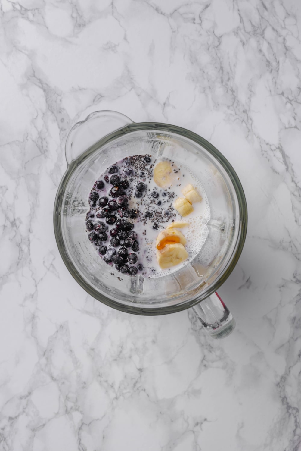 An overhead shot of a glass blender filled with milk, sliced banana, frozen blueberries, greek yogurt, and chia seeds.