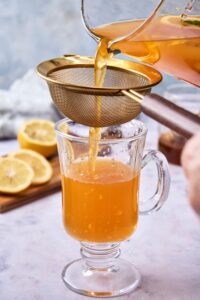 An image of flu bomb tea being poured from a glass pitcher through a small strainer into a glass tea mug.