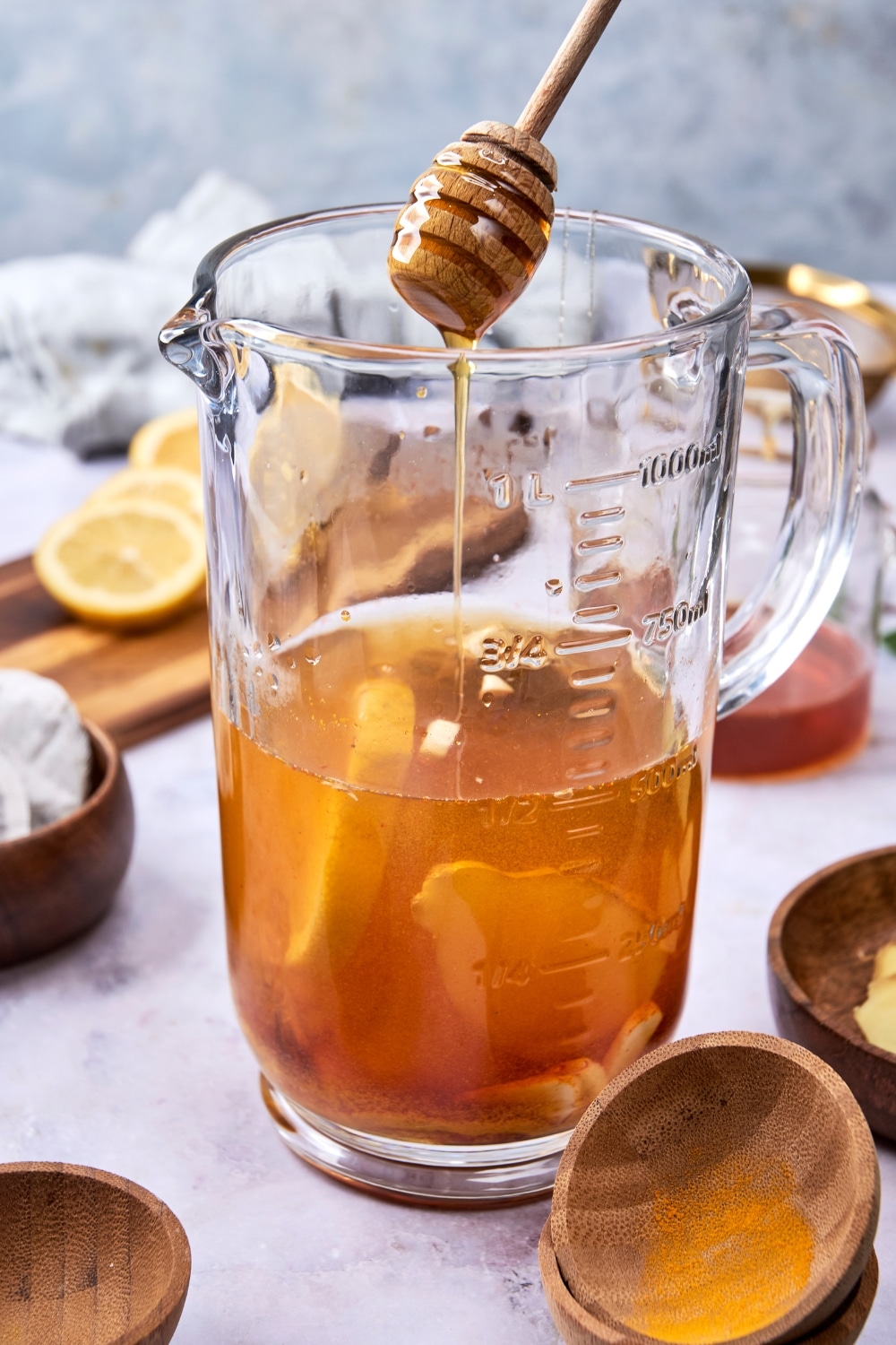 A glass pitcher filled with green tea mixture with sliced lemon and fresh ginger. There is a honey dipper pouring honey into the mixture.