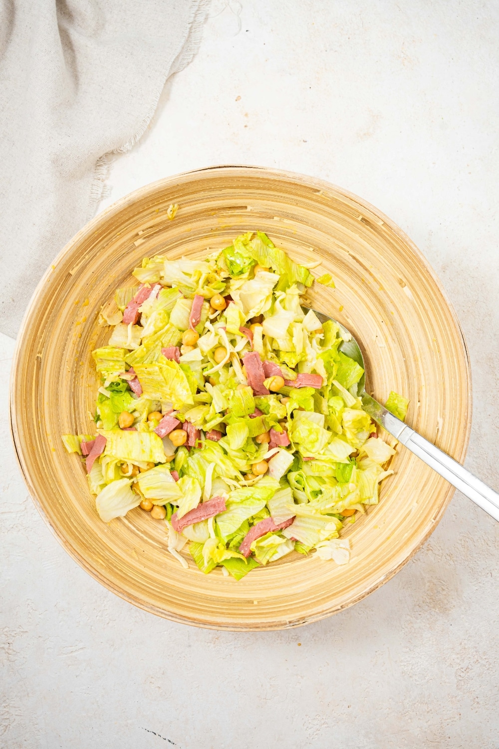 An overhead shot of a large salad bowl filled with chopped lettuce, salami, and chick peas. There is a spoon in the bowl.