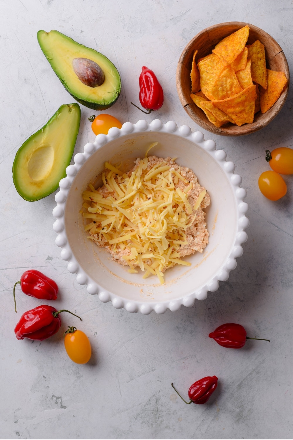 An overhead shot of a bowl of a cottage cheese queso mixture with shredded monterey jack cheese on top. There is a smaller bowl of tortilla chips and a halved avocado next to the bowl.