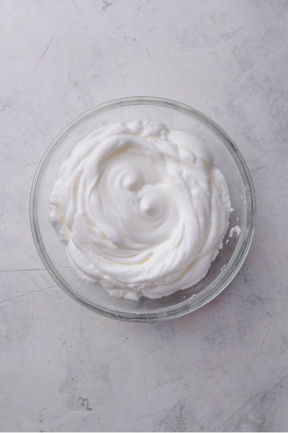 An overhead shot of a large glass bowl filled with beaten egg whites.