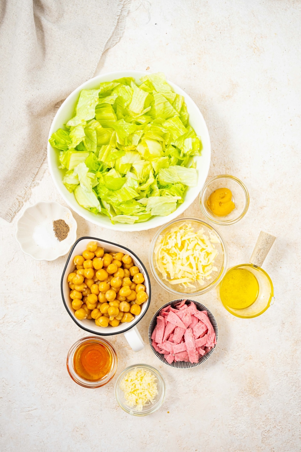 An overhead shot of several bowls of various sizes containing ingredients for the la scala chopped salad including iceberg lettuce, chick peas, salami, shredded mozzarella, olive oil, and red wine vinegar.