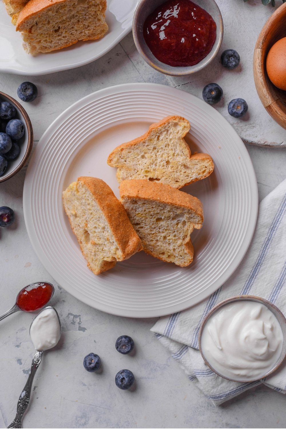 An overhead shot of a white ceramic plate with three slices of protein bread layered against one another. There is a bowl of yogurt and a bowl of jam surrounding the plate.