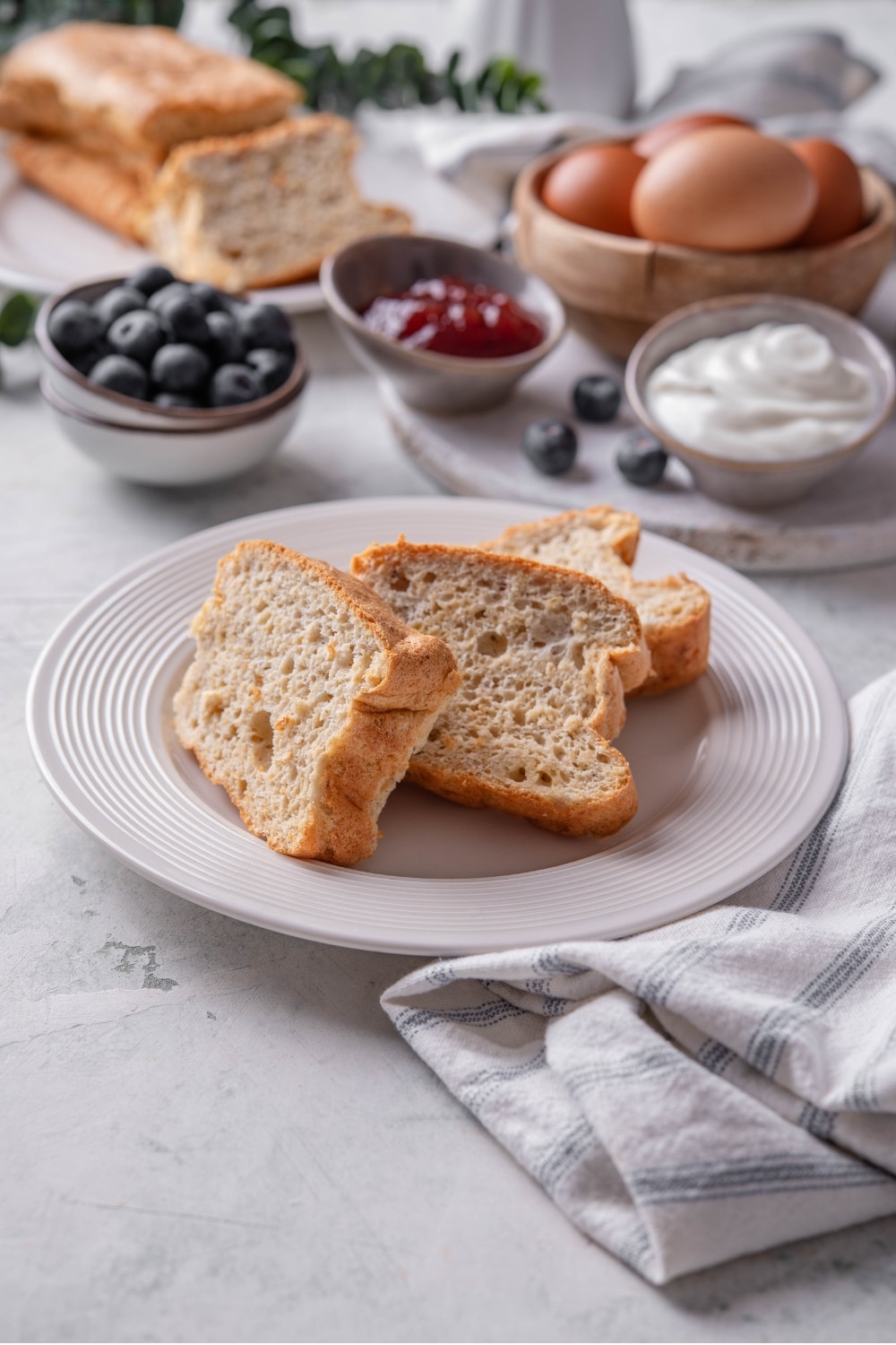 Three slices of protein bread stacked upright on a white plate. There is a napkin next to the plate and an additional loaf in the background.