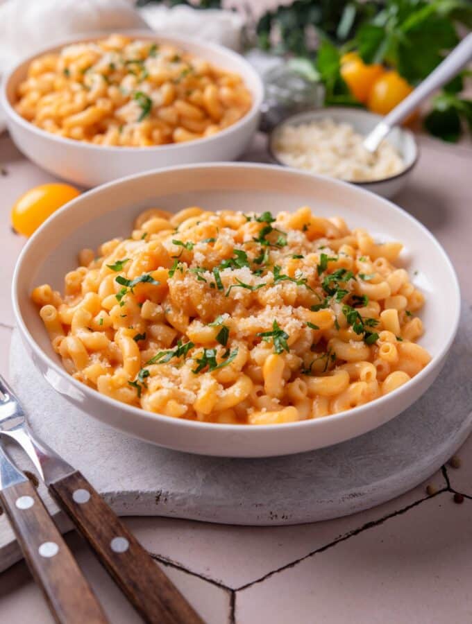 A bowl of protein mac and cheese on a white wooden server garnished with parsley and grated cheese. There are two forks next to the bowl. There is a second bowl of mac and cheese in the background.