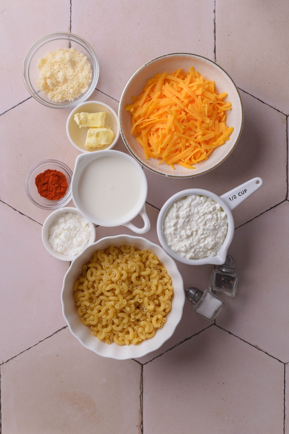 An overhead shot of several bowls containing a variety of ingredients for protein mac and cheese including elbow macaroni, shredded cheddar cheese, cottage cheese, milk, butter, and corn starch.