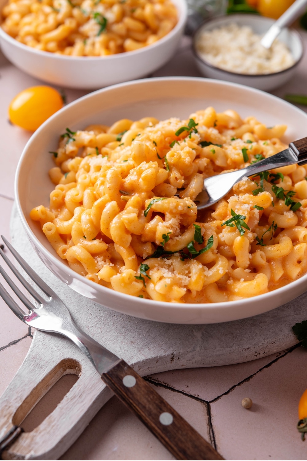 A bowl of protein mac and cheese on a white wooden server. There is a fork inserted into the macaroni and another fork next to the bowl. There is a second bowl of mac and cheese in the background.