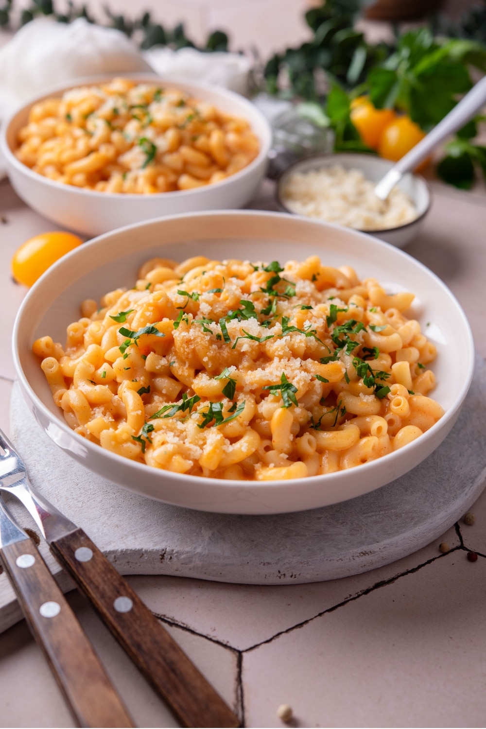 A bowl of protein mac and cheese on a white wooden server garnished with parsley and grated cheese. There are two forks next to the bowl. There is a second bowl of mac and cheese in the background.