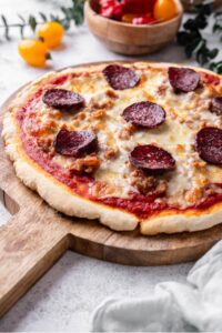 A whole unsliced protein pizza on a wooden server. The pizza is topped with cheese, pepperoni, and sausage.