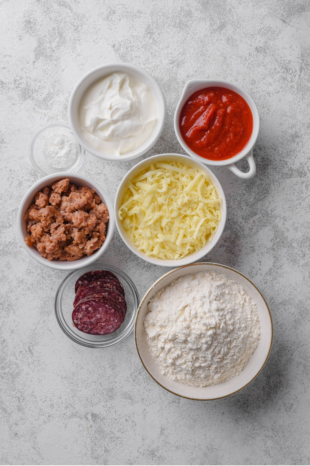 An overhead shot of several bowls holding various ingredients to make protein pizza including flour, shredded mozzarella cheese, pizza sauce, crumbled sausage, sliced pepperoni, and greek yogurt.