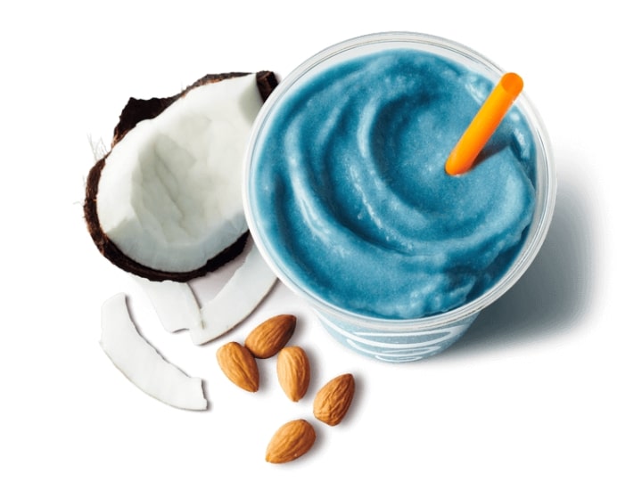 A blue smoothie in a cup with a coconut and almonds next to it.