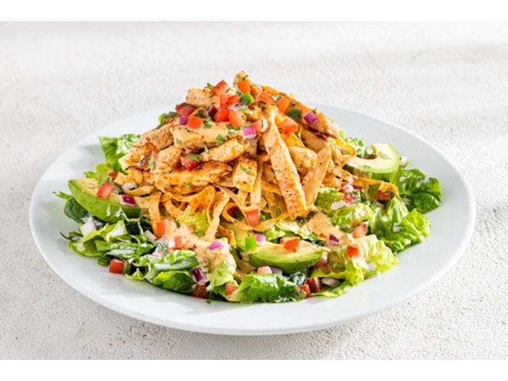 Grilled chicken on top of a mixed greens salad with avocado and diced tomato in a white bowl.