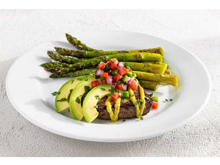 A bunch of avocado slices on top of a piece of steak with grilled asparagus on a white plate.